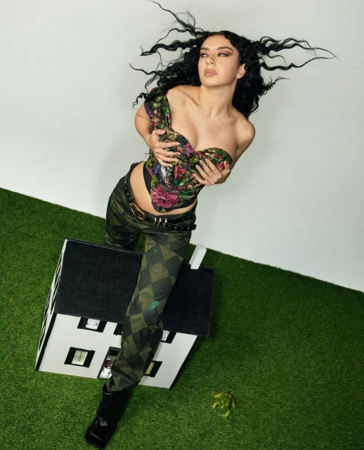 Portrait of a woman in a floral corset top, patterned pants, and black boots posing with a white dollhouse on artificial turf.