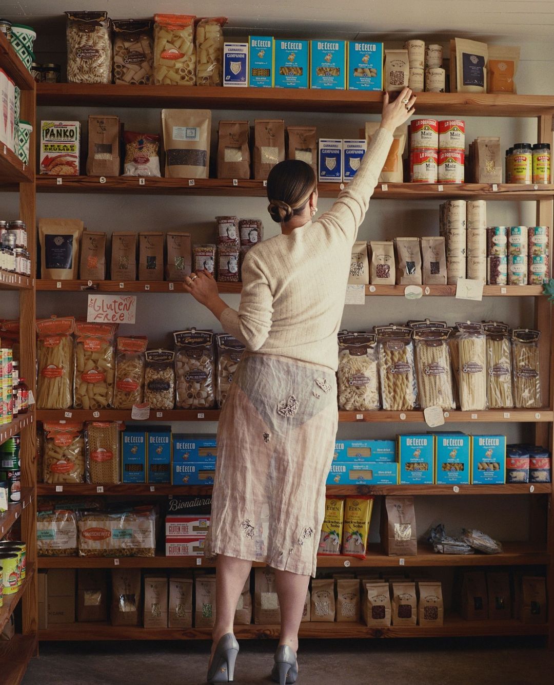 Woman in skirt reaches for pasta on rustic wooden shelves in small grocery store with bags and boxes of various dry goods.
