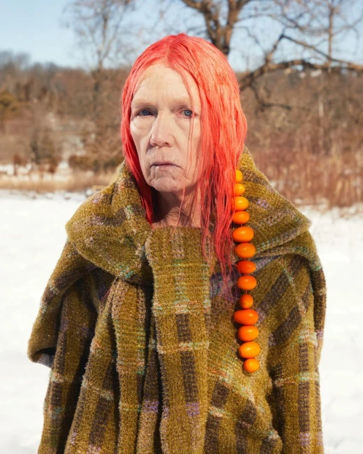 Older adult woman with red hair and amber bead strand poses in a winter field wearing a plaid, tweed coat, creating a striking portrait.
