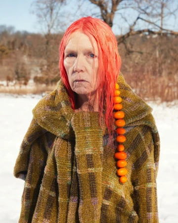 Older adult woman with red hair and amber bead strand poses in a winter field wearing a plaid, tweed coat, creating a striking portrait.