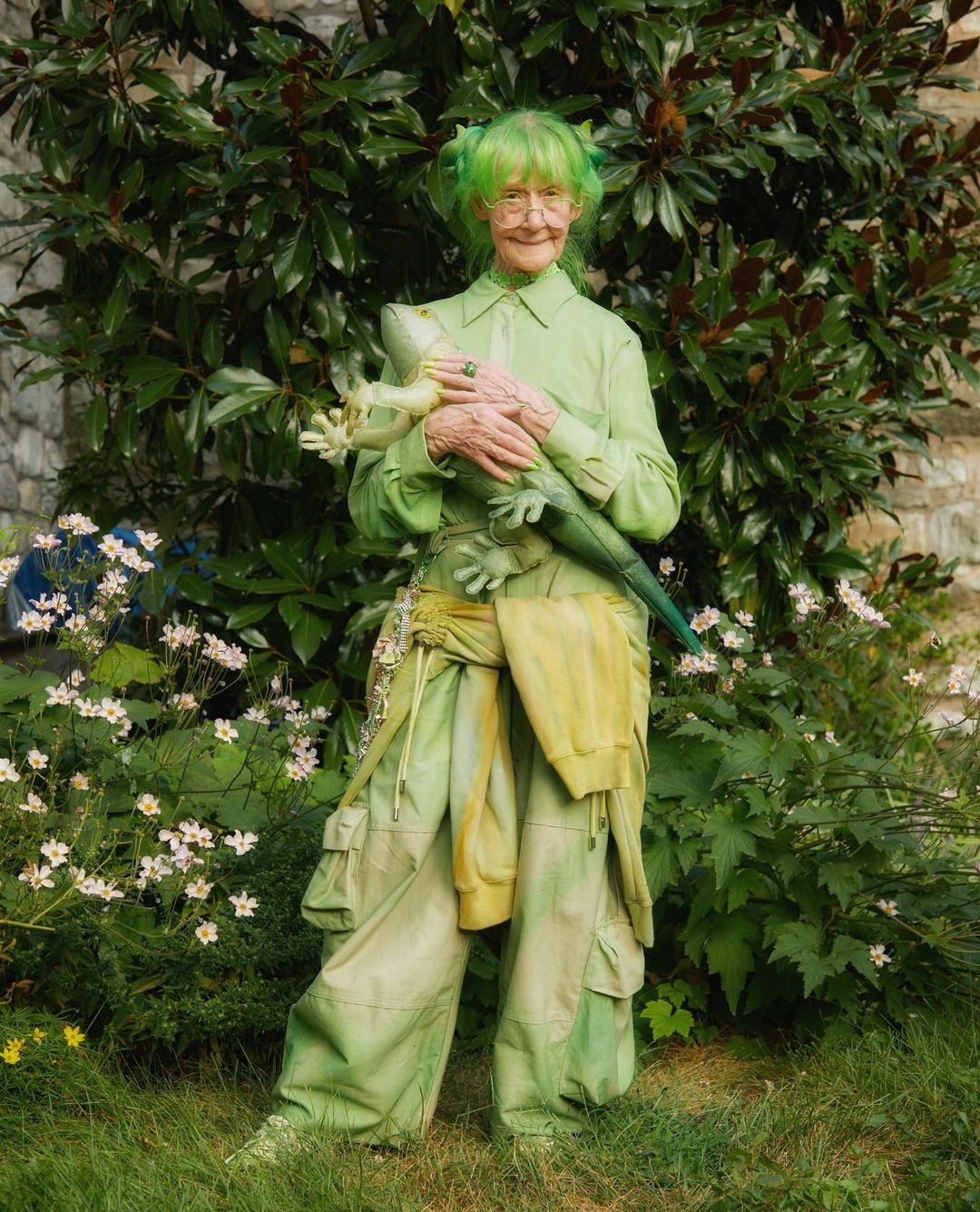 Elderly woman with bright green hair and a green outfit poses with a stuffed lizard in a lush, green outdoor setting near white flowers.
