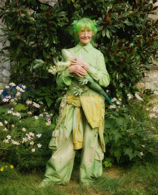 Elderly woman with bright green hair and a green outfit poses with a stuffed lizard in a lush, green outdoor setting near white flowers.