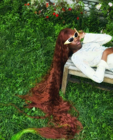 Black woman with extremely long auburn hair wearing sunglasses lying on a wooden bench outdoors in tall green grass by Charlie Engman