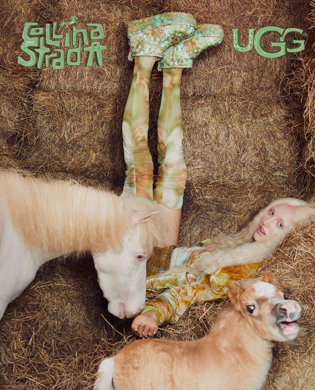 Blonde woman in patterned outfit lying in hay with two ponies, floral UGG boots, and Stella Studios logo in an indoor stable.