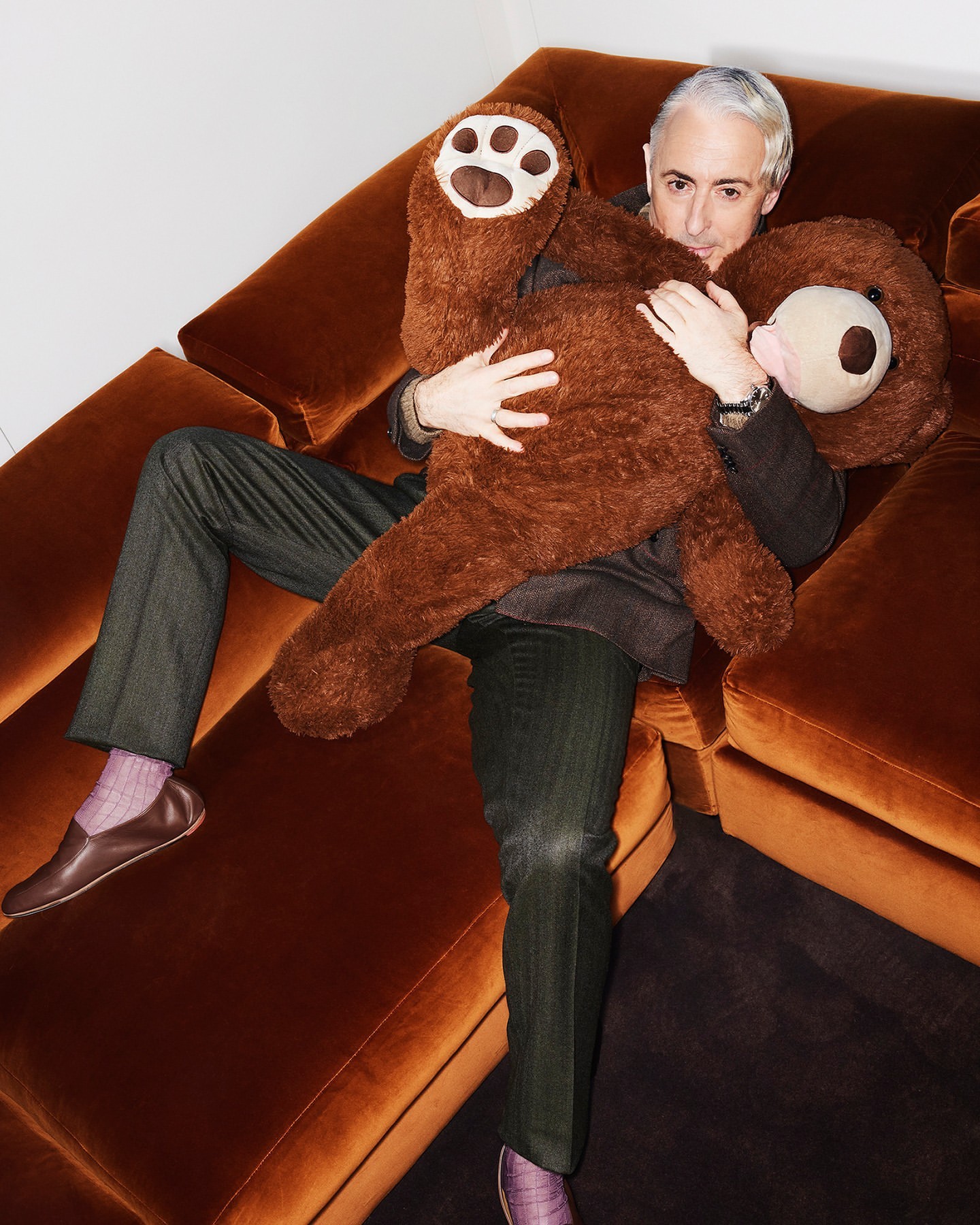 Man on an orange couch hugs a large brown teddy bear, wearing brown slacks, a blazer, and brown leather slippers. Indoor setting.