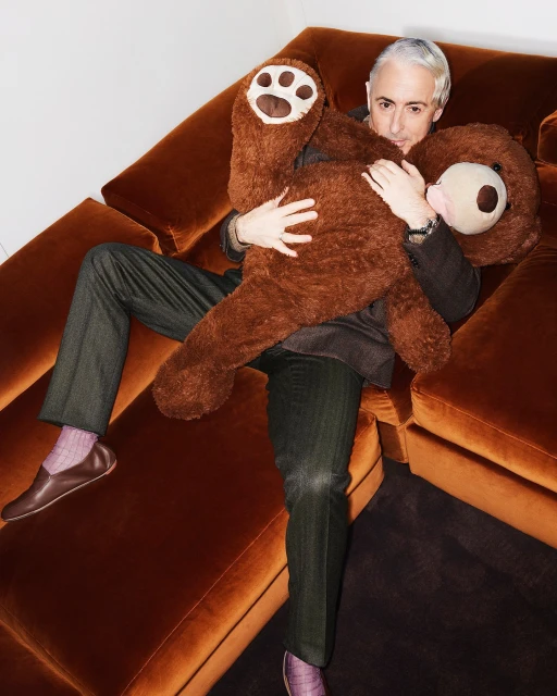 Man on an orange couch hugs a large brown teddy bear, wearing brown slacks, a blazer, and brown leather slippers. Indoor setting.