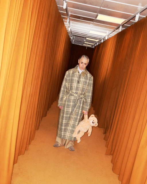Adult male in a plaid coat stands in an orange hallway holding a teddy bear, with indoor lighting and a straight-on, medium shot.