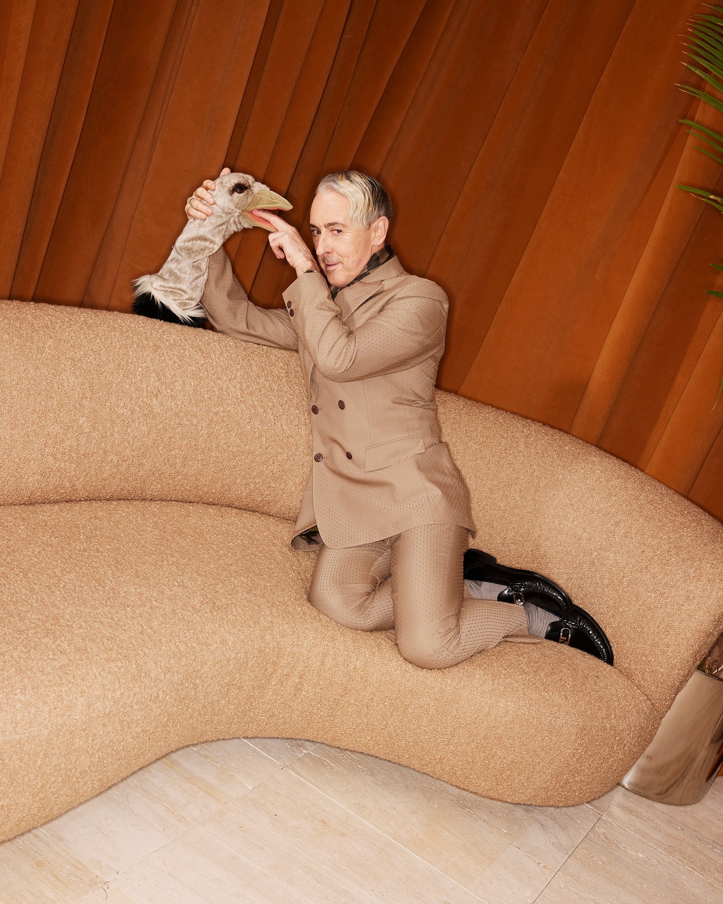 Man in a tan suit poses on a beige couch, holding a bird puppet toward the camera in an indoor setting with a brown curtain.