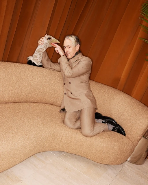 Man in a tan suit poses on a beige couch, holding a bird puppet toward the camera in an indoor setting with a brown curtain.
