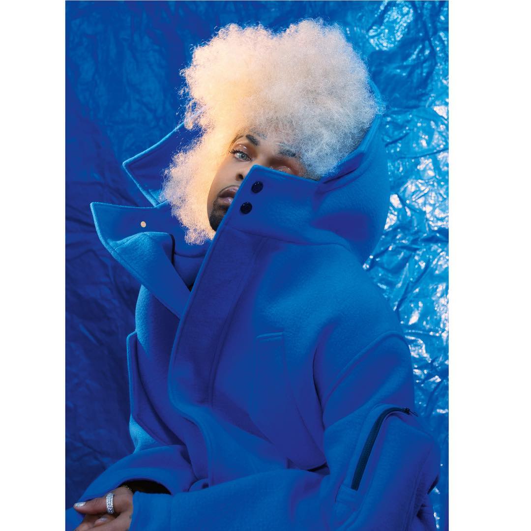 Man with platinum afro wearing a blue coat poses against a matching blue background in a striking portrait. Fashion, style, and portraiture.
