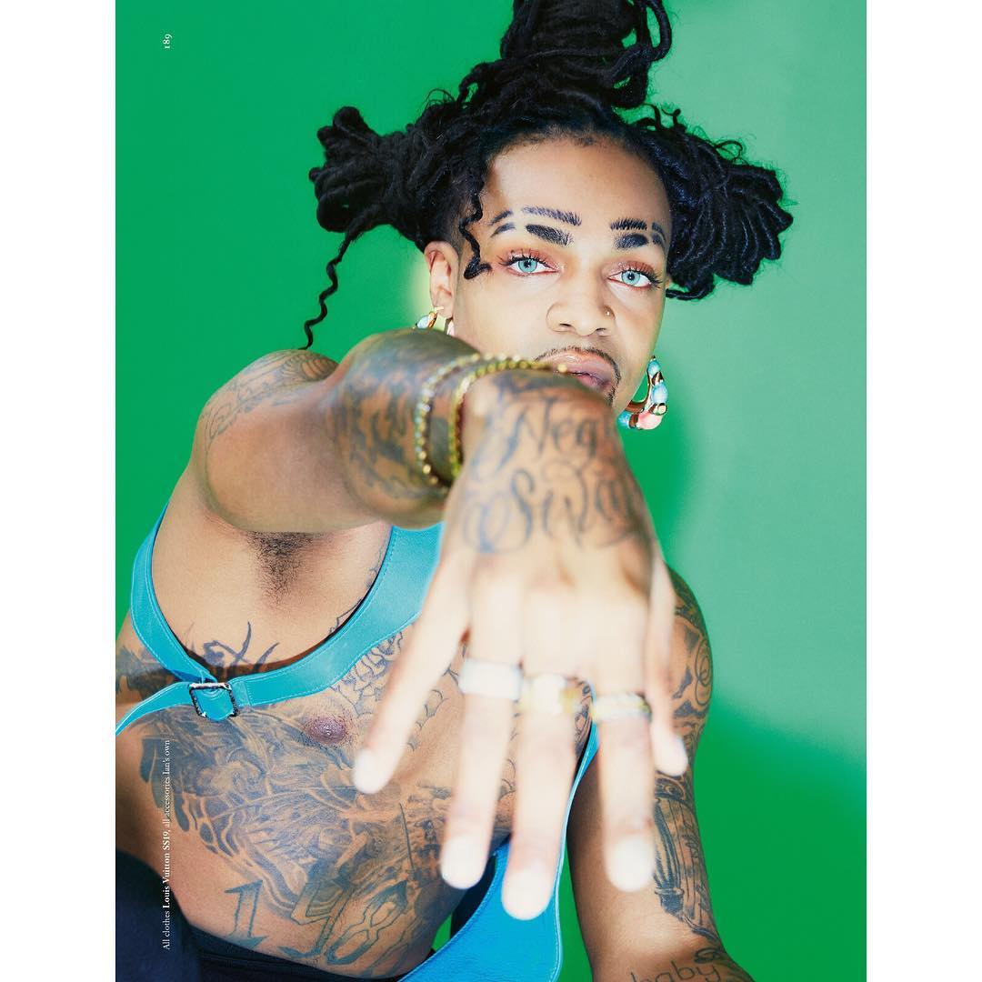Portrait of a non-binary person with locs, tattoos, and blue eyes, wearing a teal harness, posing in front of a vibrant green backdrop.