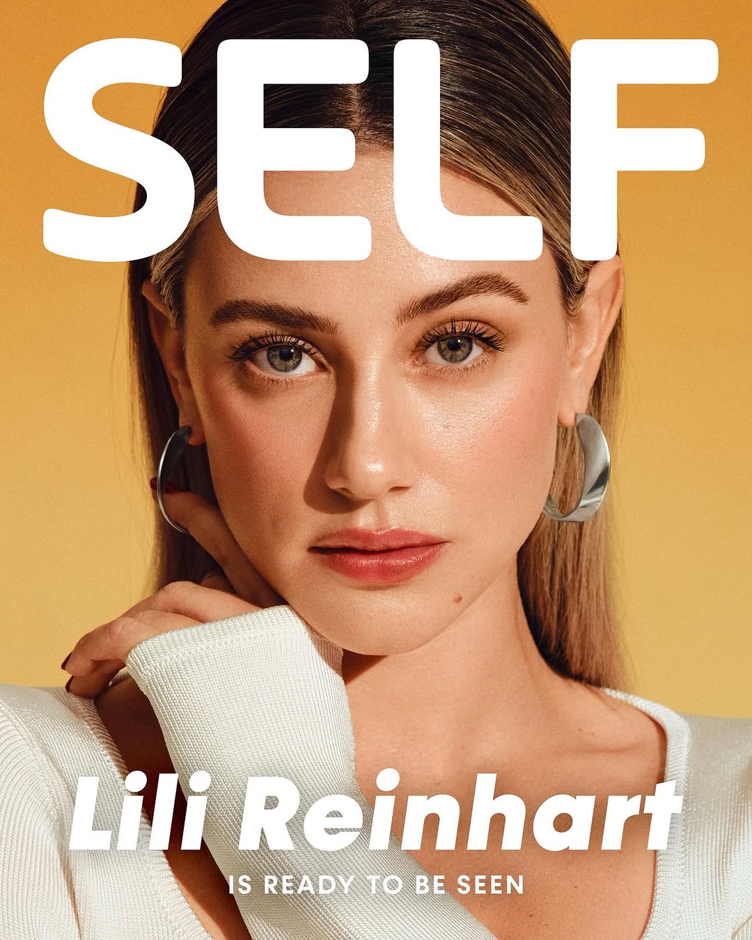 Close-up portrait of Lili Reinhart on a yellow background, wearing a white sweater and silver hoop earrings for magazine cover.