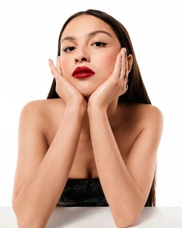Close-up portrait of Olivia Rodrigo with bold red lipstick and hands framing her face against a white background