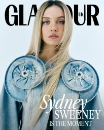 Sydney Sweeney with long blonde hair and freckles wearing a blue denim top with large circular shoulder appliqués against a white backdrop.
