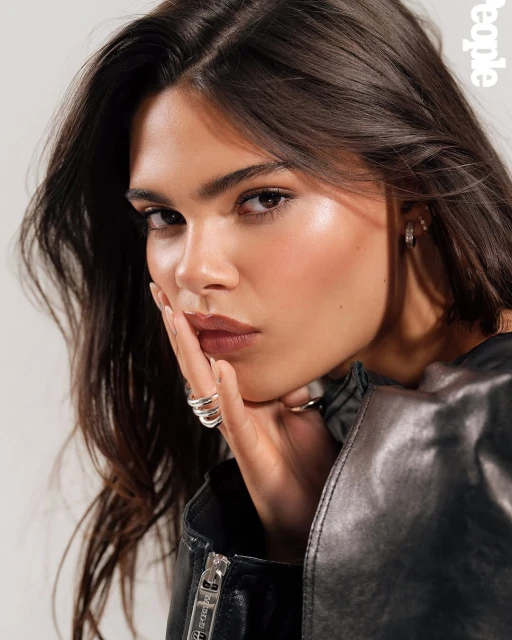 Close-up portrait of a woman with dark hair in a black leather jacket against a white backdrop, gazing directly into the camera.