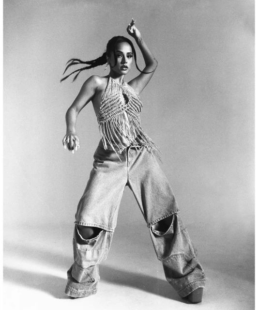 A striking portrait of a woman in a halter top and ripped jeans strikes a dance pose against a neutral backdrop in black and white.