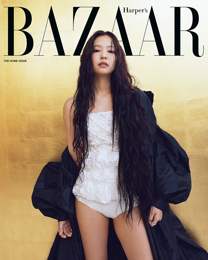 A woman with long dark wavy hair wearing a white strapless dress and a black coat against a gold wall
