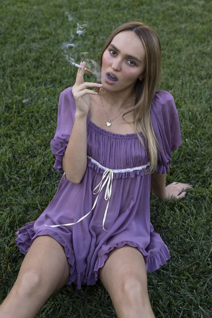 Young woman sits on grass wearing a purple dress, smoking a cigarette with visible smoke around her face, in an outdoor setting.