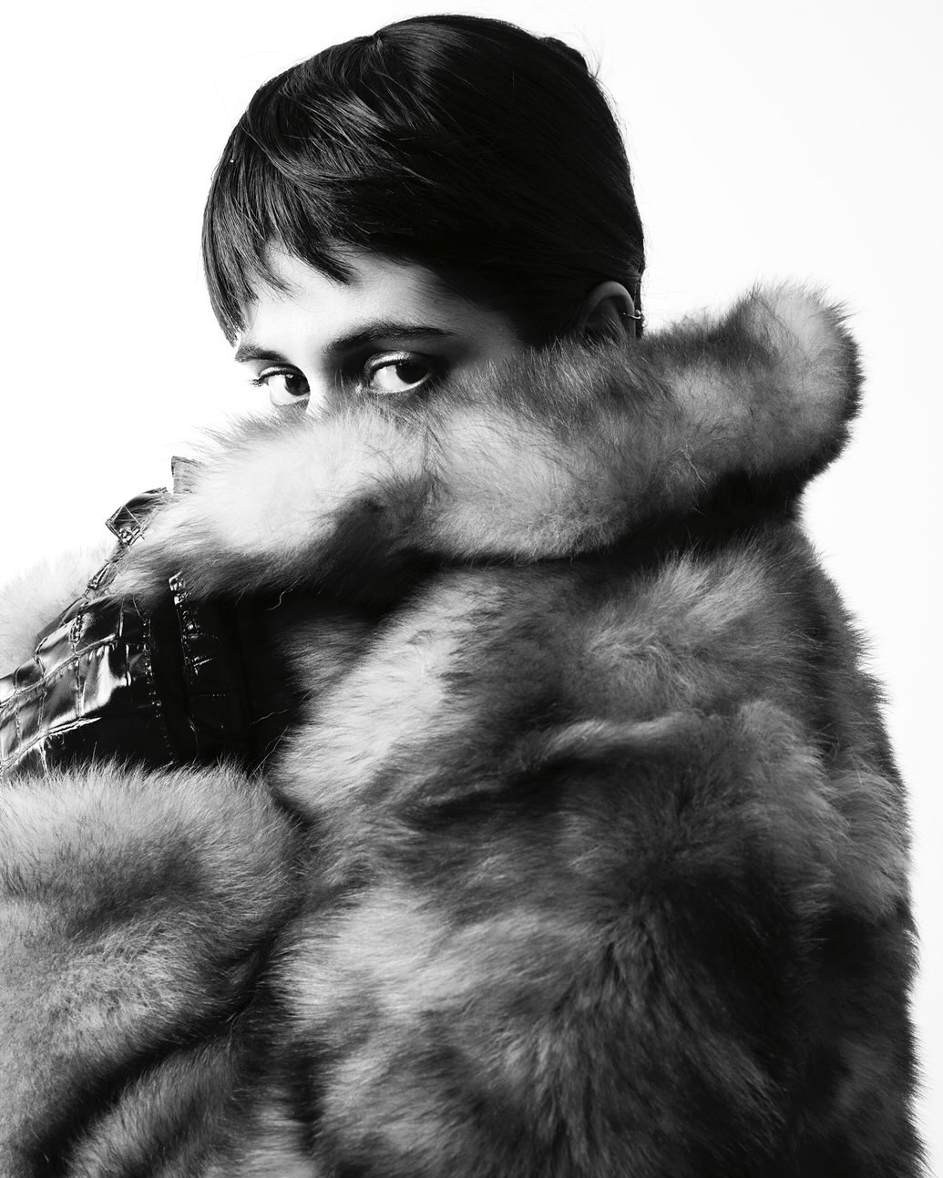 Monochrome portrait of a young adult woman with a fur coat, looking at the camera against a white background in a studio setting.