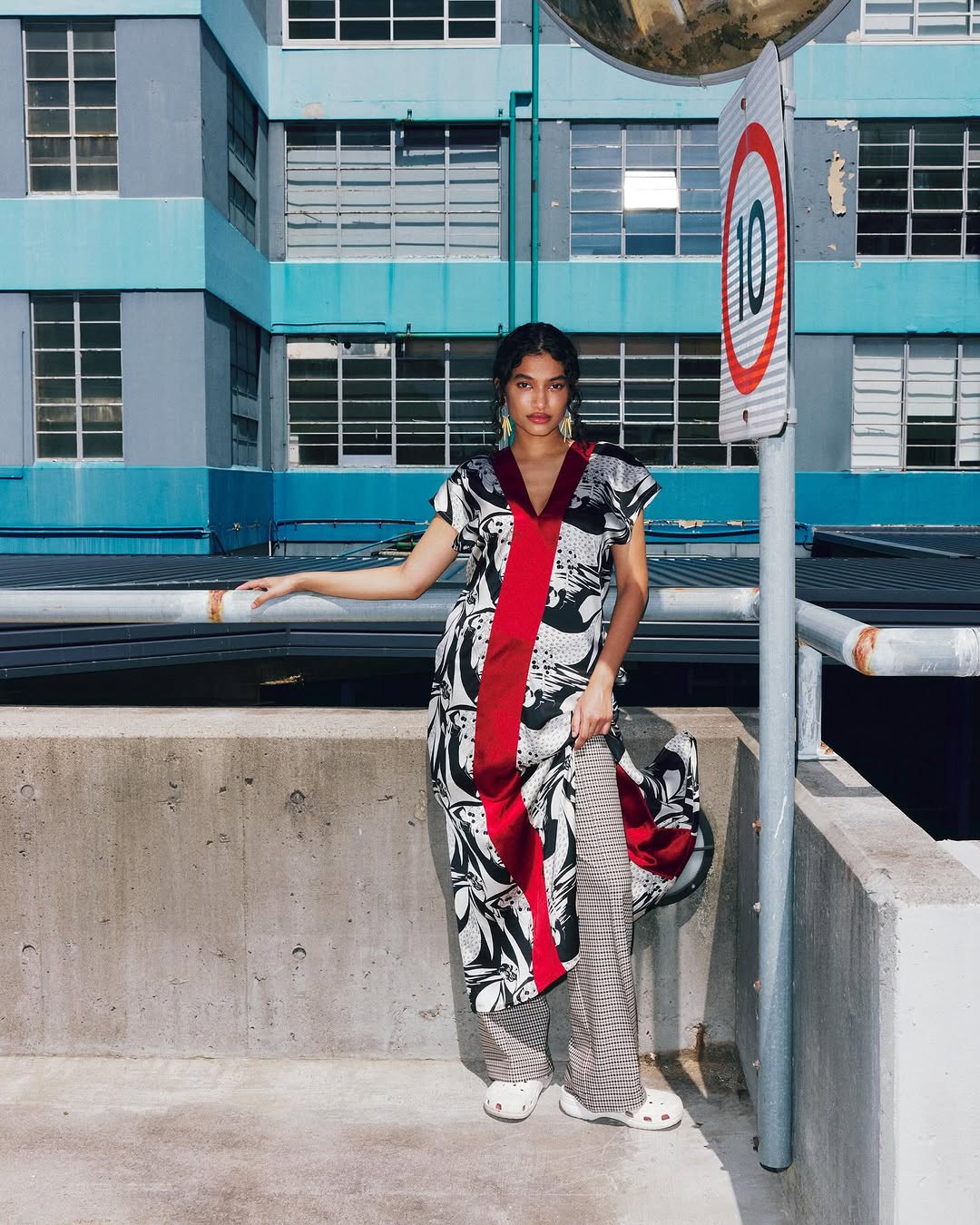 Woman poses in a modern dress with a red stripe on an urban rooftop next to a street sign, showcasing fashion and architecture.