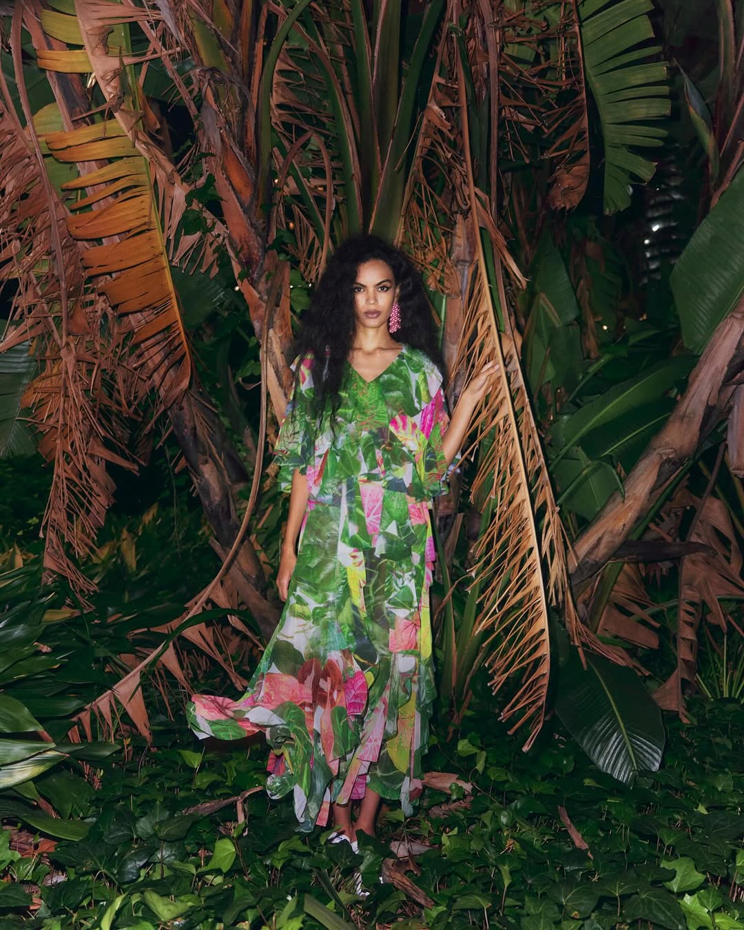 A woman with dark curly hair poses in a green floral dress among lush tropical foliage and palms in an outdoor setting.