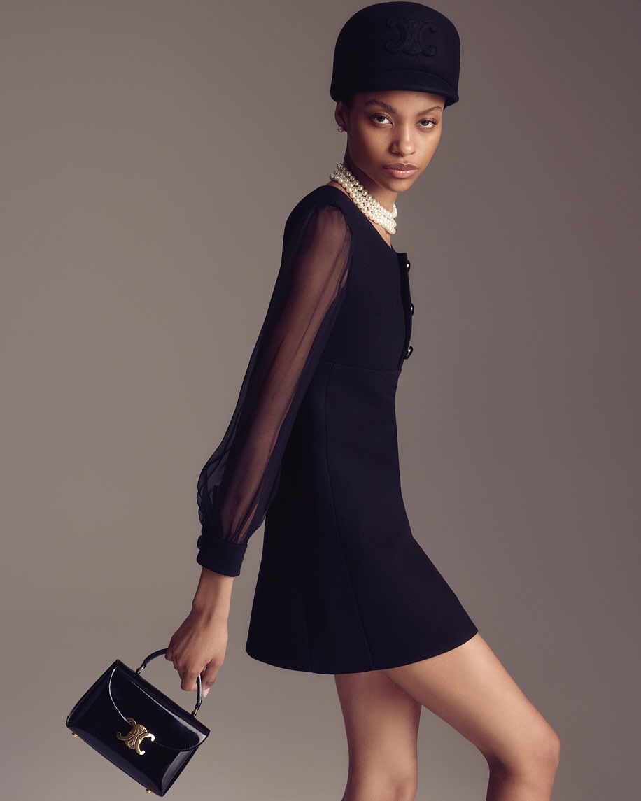Elegant Black Fashion: A poised woman in a black dress, cap, and pearl necklace holds a black purse against a neutral backdrop.