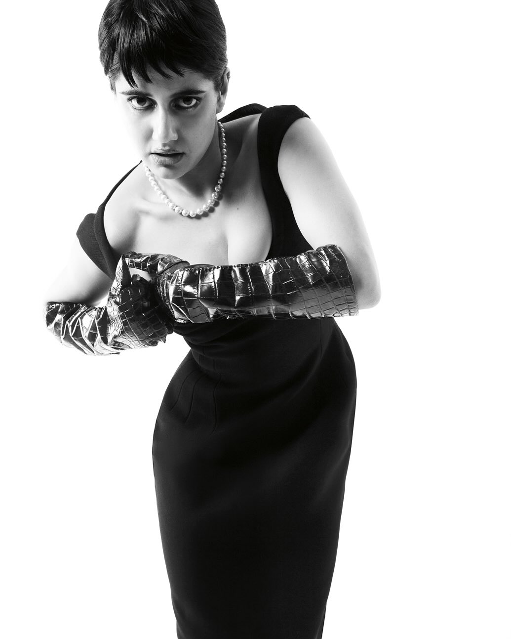 Black and white portrait of a woman in a fitted black dress and shiny, textured gloves, looking directly at the camera with a neutral expression.