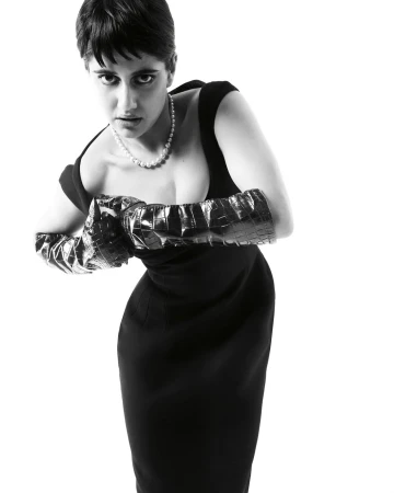 Black and white portrait of a woman in a fitted black dress and shiny, textured gloves, looking directly at the camera with a neutral expression.