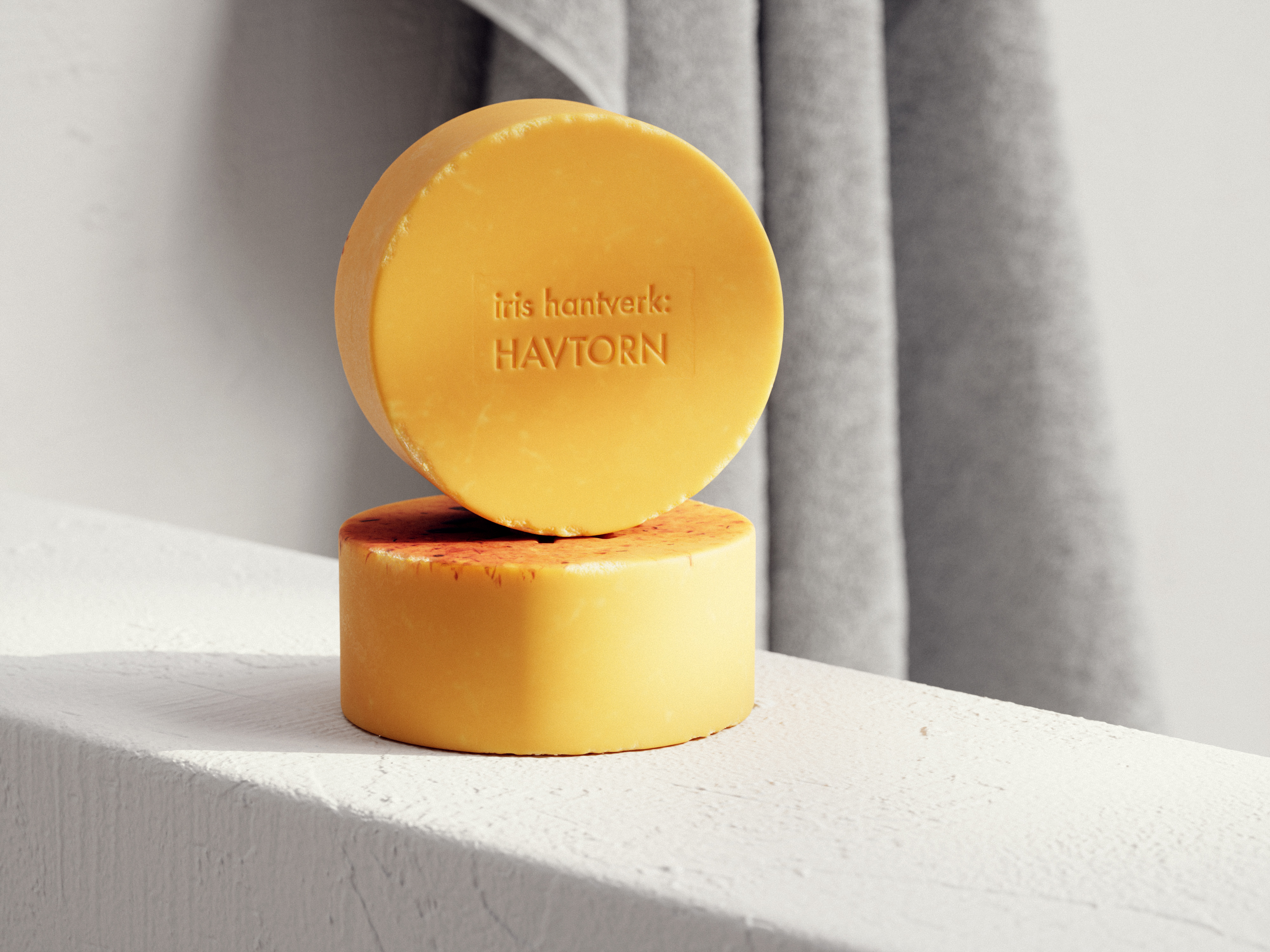 Two round bars of yellow soap stacked on a white surface against a light background with a grey towel partially visible.