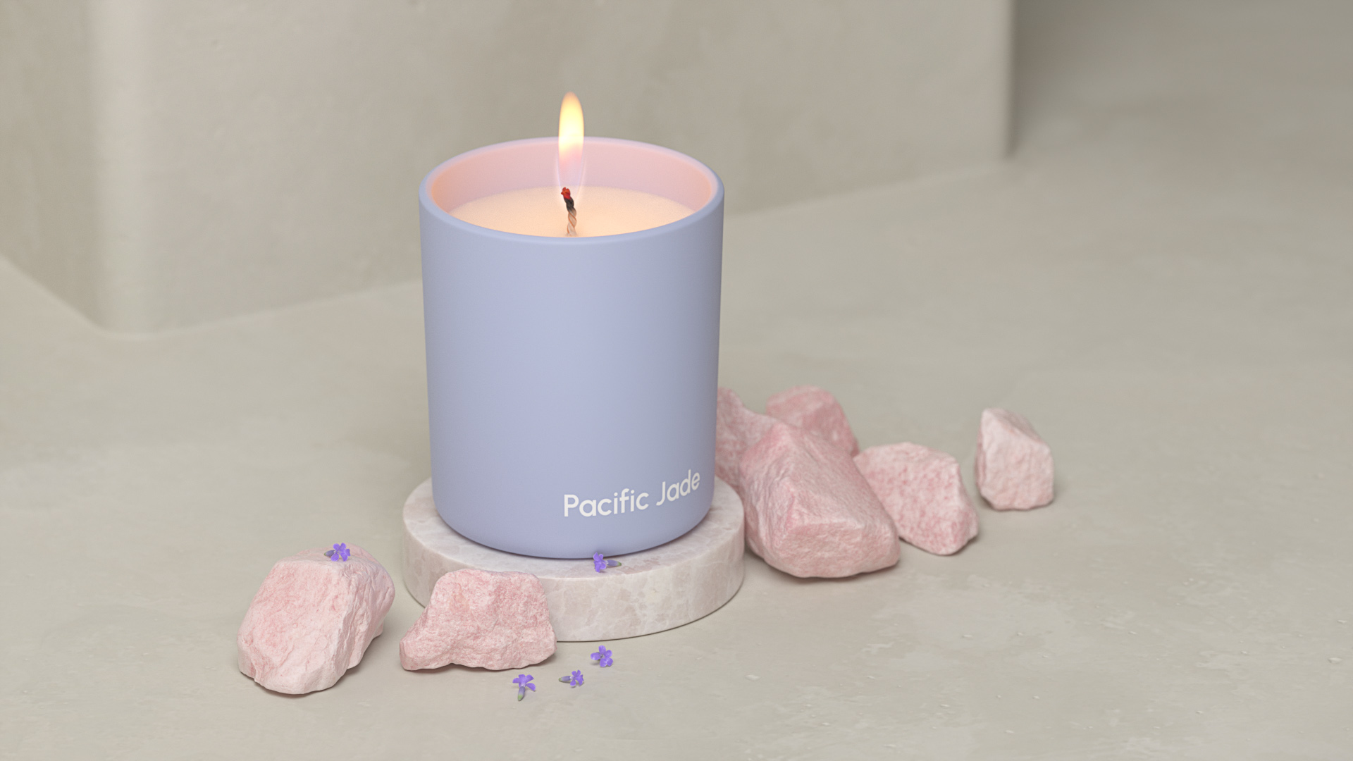Lit candle in a periwinkle container with pink accents surrounded by small pink rocks and lavender flowers against a gray backdrop.