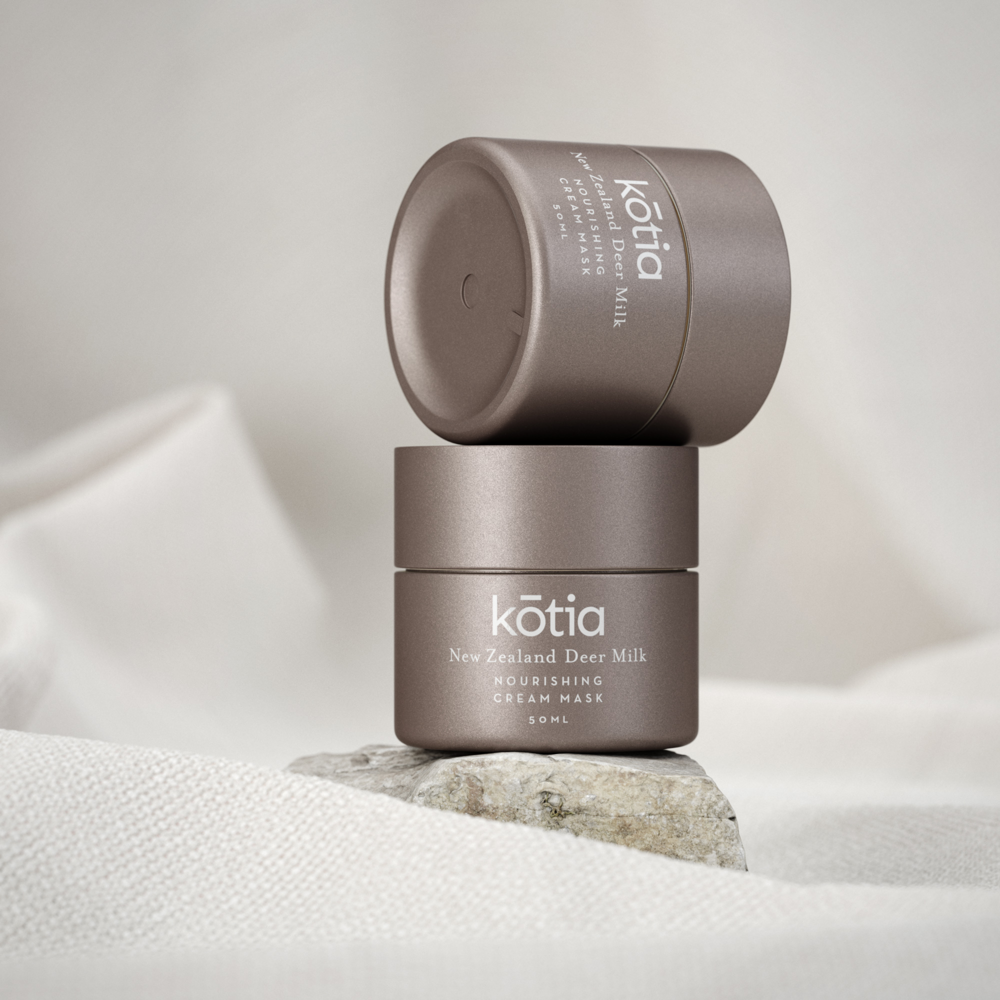 Two jars of Kōtia New Zealand Deer Milk Nourishing Cream Mask are stacked on a rock with a soft, folded white cloth backdrop.