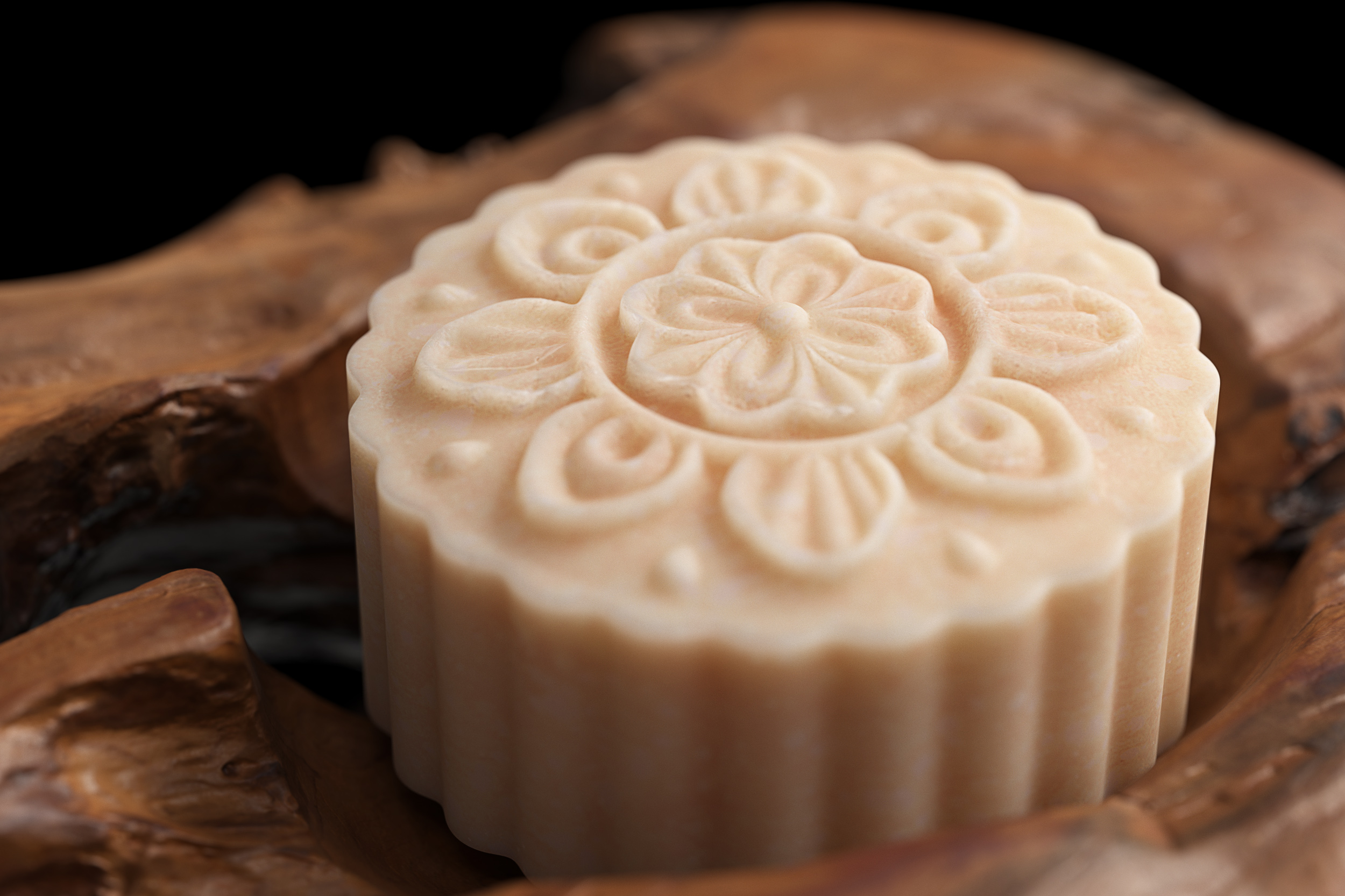 Close-up of ornate, light-beige mooncake on a textured wood surface against a black backdrop. Food, baked good, pattern detail.