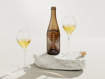 Brown glass wine bottle labeled Pernelle with two stemmed wine glasses filled with white wine resting on a grey stone block.