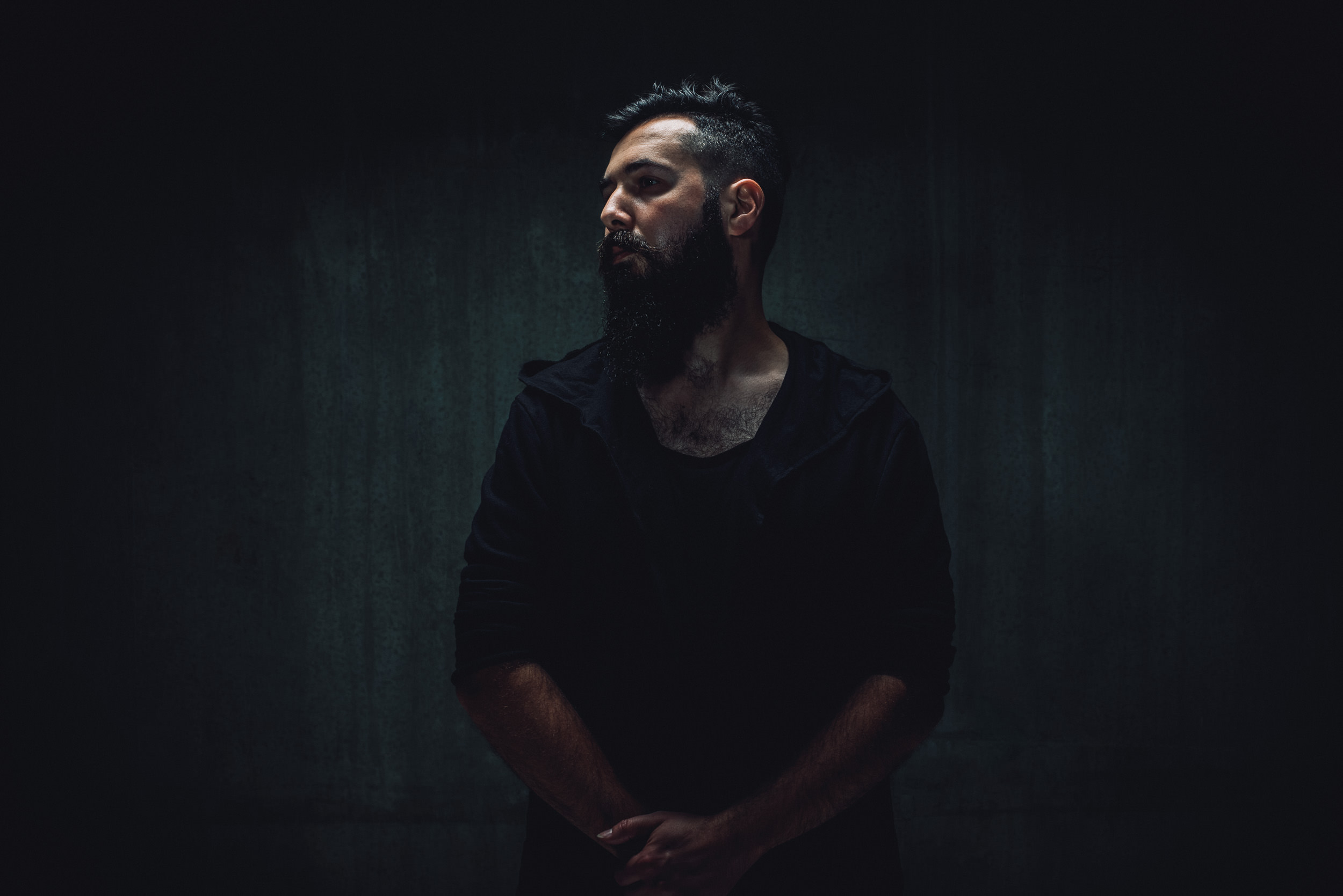 Portrait of a bearded man in a hooded shirt against a dark gray wall, captured in a dimly lit, moody atmosphere.