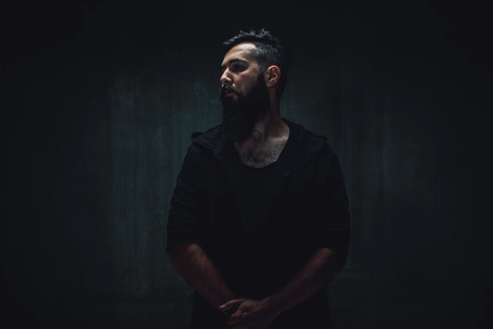 Portrait of a bearded man in a hooded shirt against a dark gray wall, captured in a dimly lit, moody atmosphere.