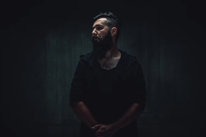 Portrait of a bearded man in a hooded shirt against a dark gray wall, captured in a dimly lit, moody atmosphere.