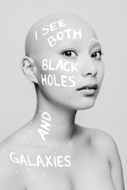 Striking black and white portrait of a bald woman with text art on her face and body, posing in a photo studio with neutral background.