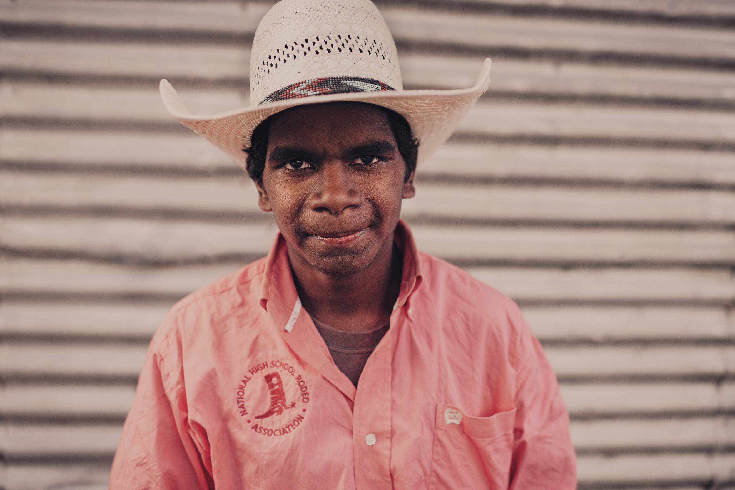 Portrait of a person wearing a straw cowboy hat and pink shirt with a logo. The person is in front of a wall with horizontal lines.