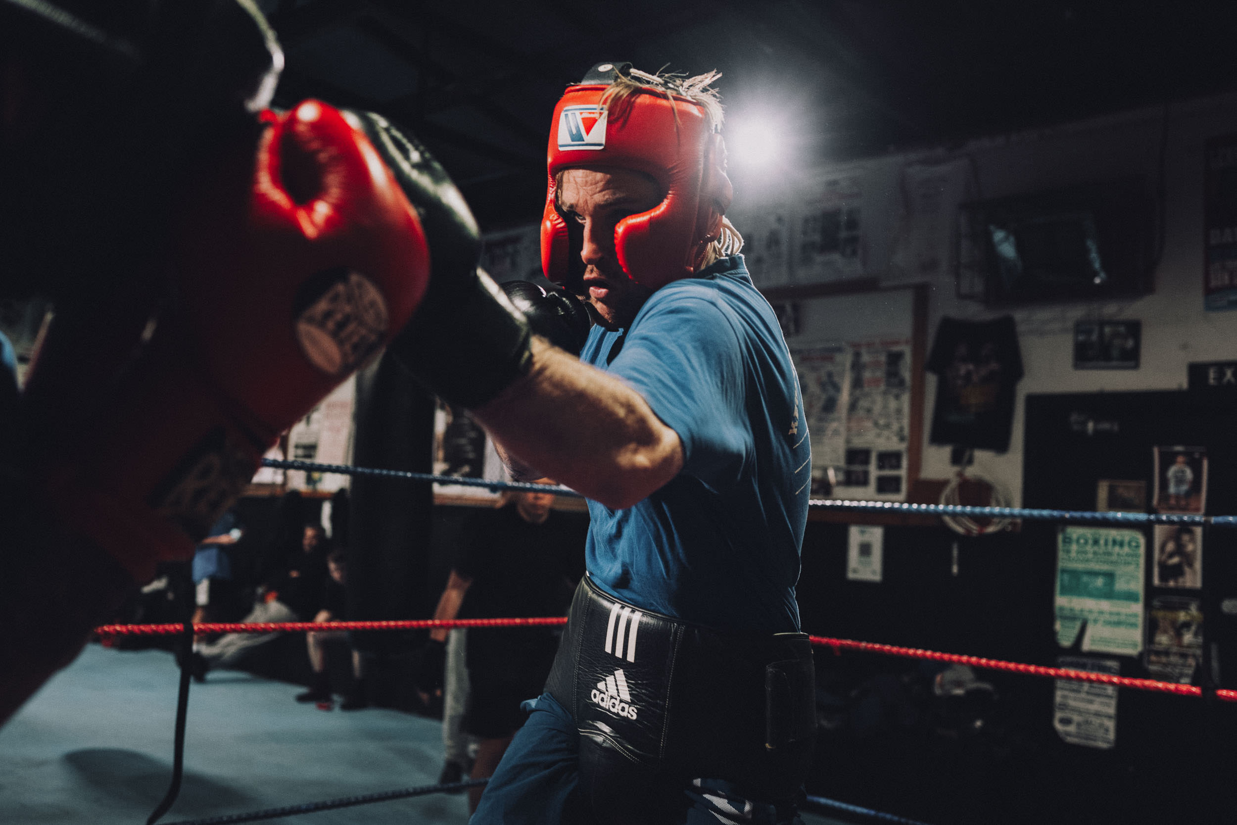 Two boxers in a boxing ring, one throwing a punch toward the other who is wearing a red head guard, Adidas athletic tape.