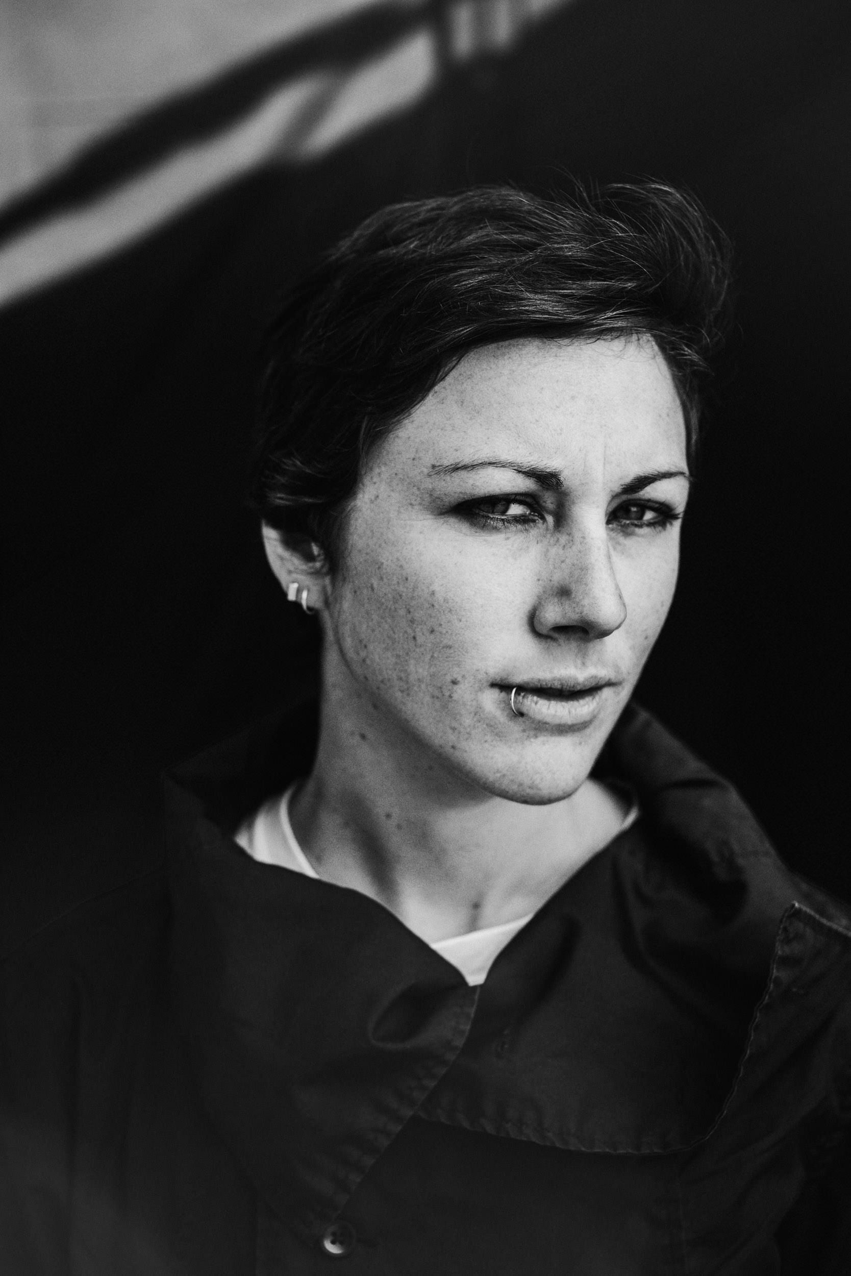 Monochrome portrait of an adult woman with short dark hair, silver lip ring, and hoop earrings against a dark shadowed background.