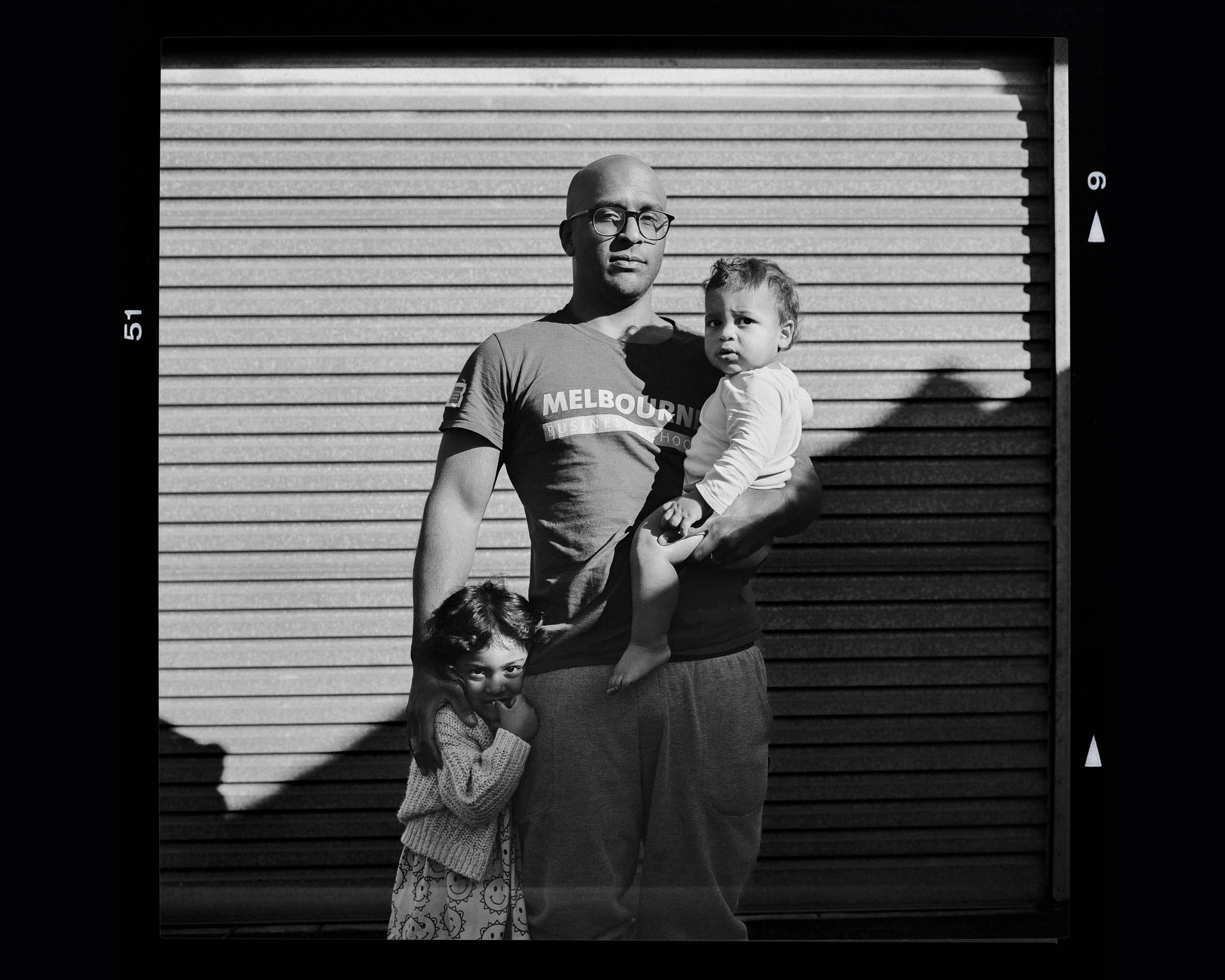 Black and white portrait of a bald man with glasses holding two young children in front of a closed metal shutter door.