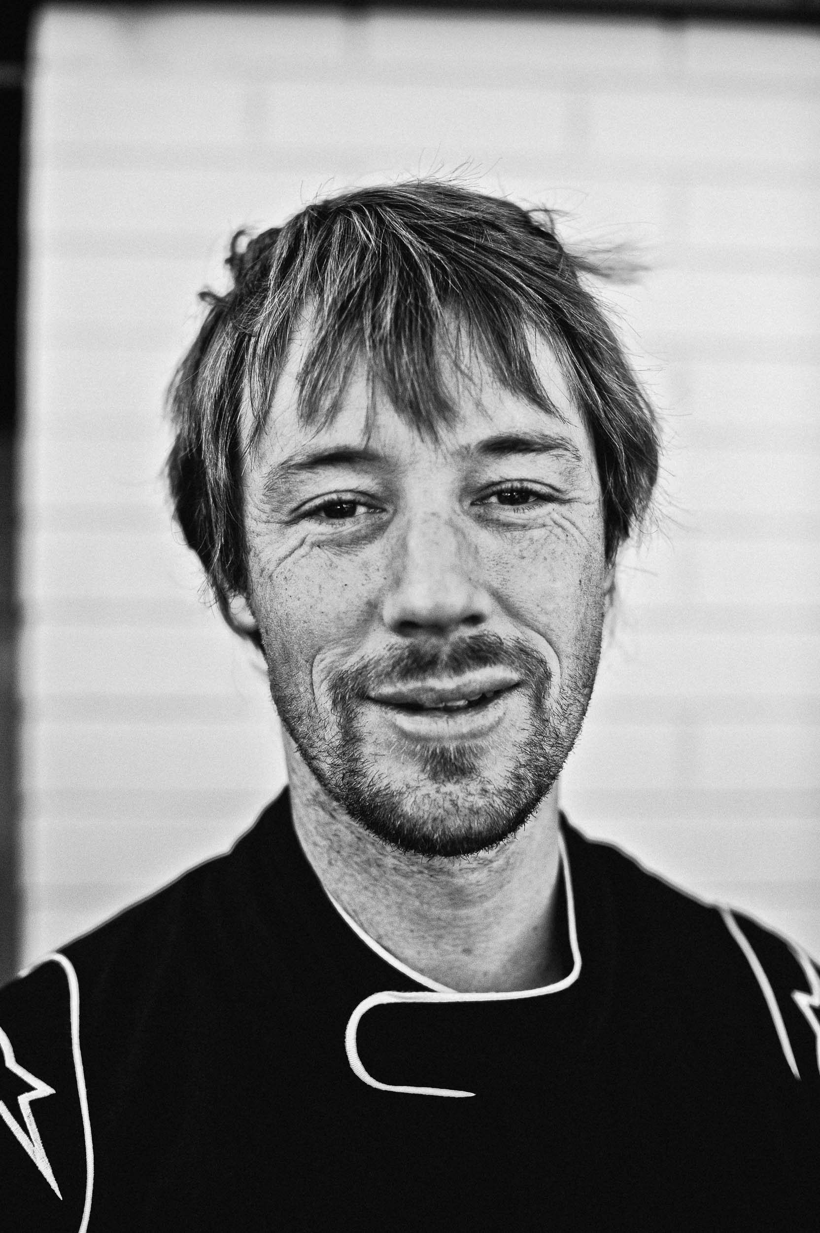 Monochrome portrait of a man with short hair and stubble wearing a racing suit against a white brick wall in Melbourne.