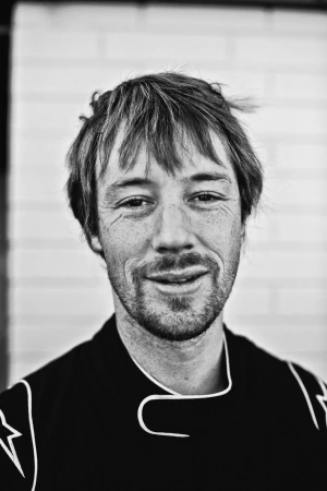 Monochrome portrait of a man with short hair and stubble wearing a racing suit against a white brick wall in Melbourne.