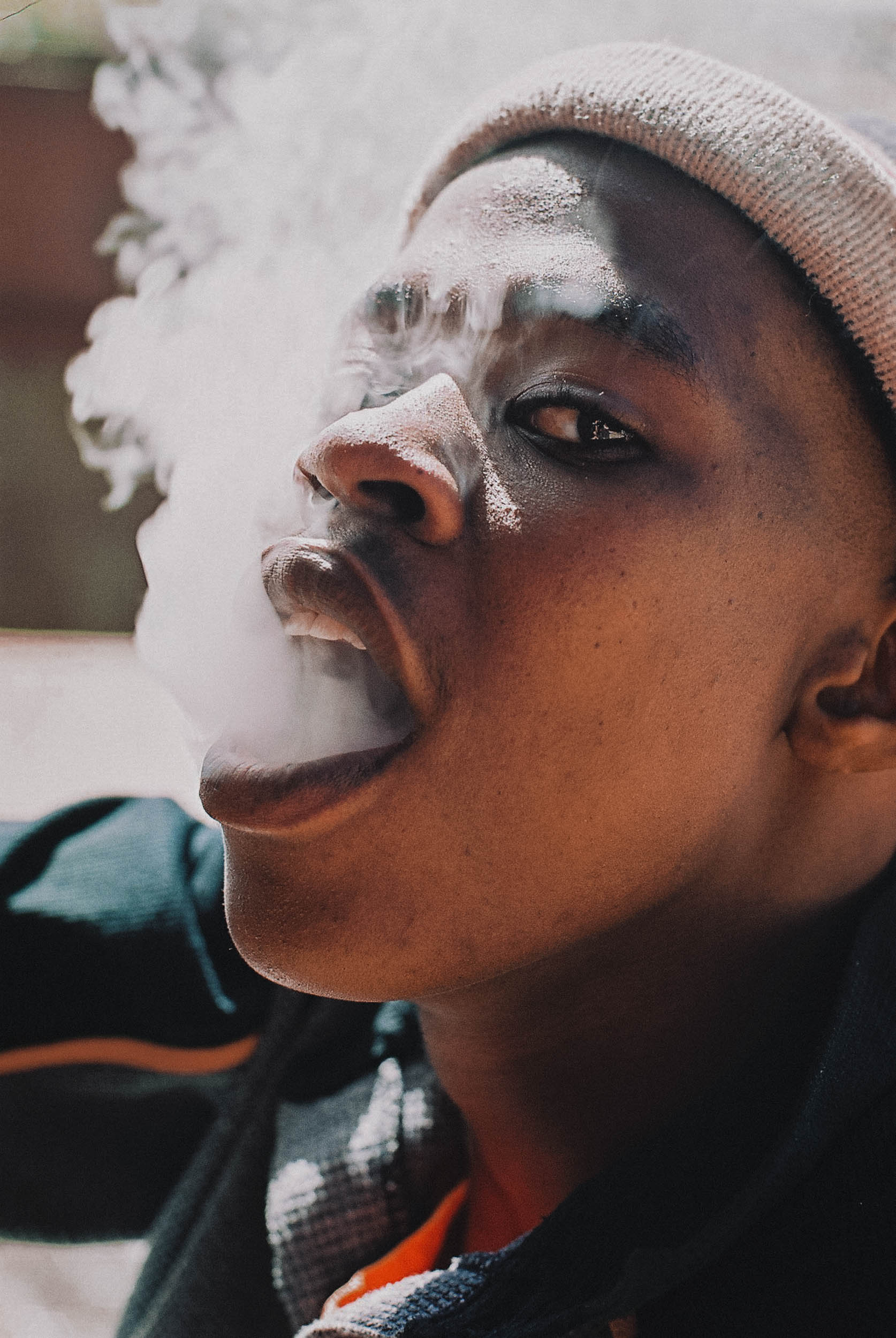 Close-up of a young man with dark skin wearing a beanie, exhaling a cloud of vapor or smoke. Shot in soft, diffused lighting.