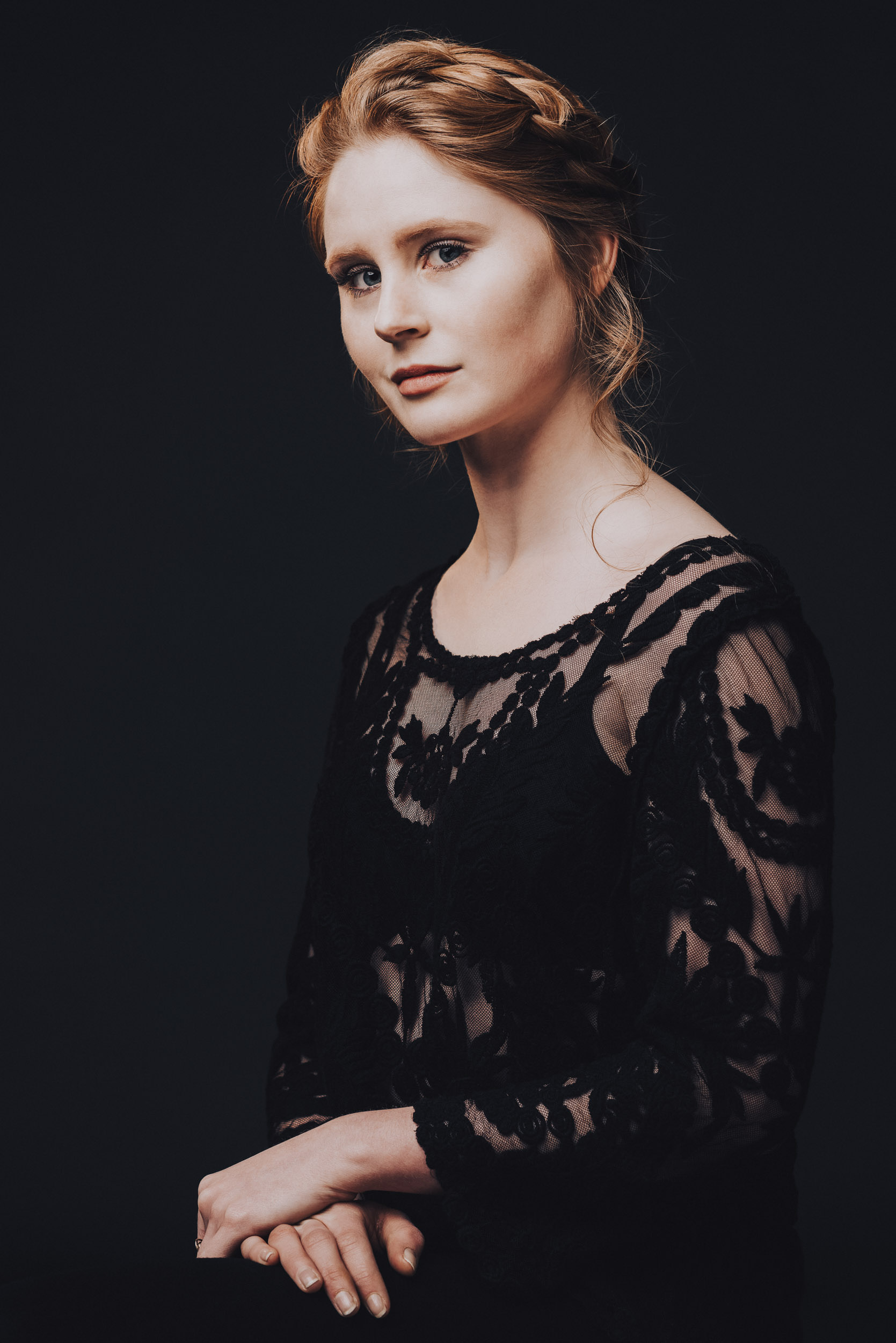 Portrait of a red-haired woman in a black lace dress against a dark background, with her hands clasped in front of her.