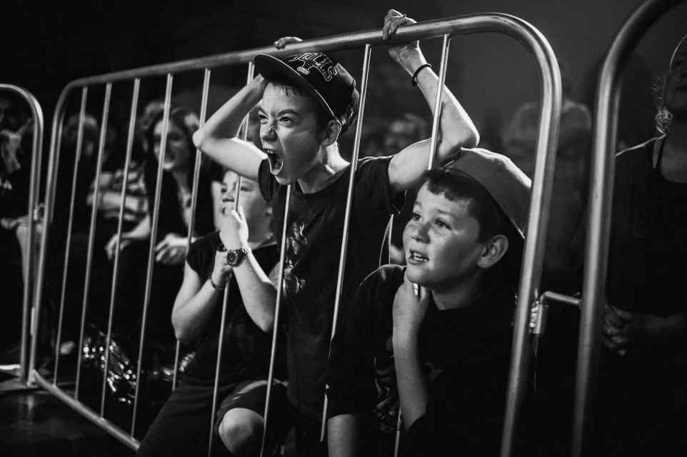Black and white shot of a group of excited children watching an event behind a metal barrier, with one child shouting enthusiastically.