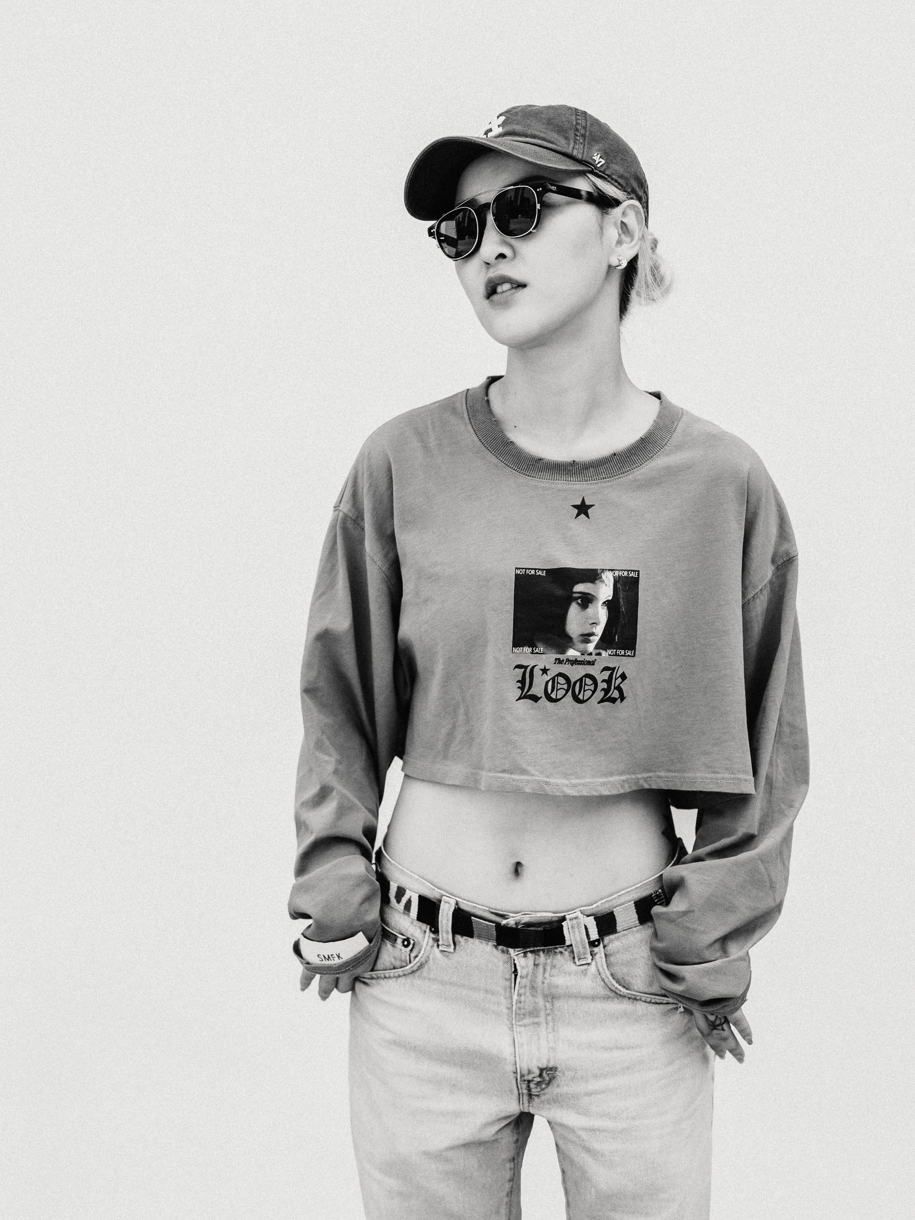 Monochrome portrait of a stylish young woman wearing a baseball cap, sunglasses, and a crop top with a graphic print, against a white background.