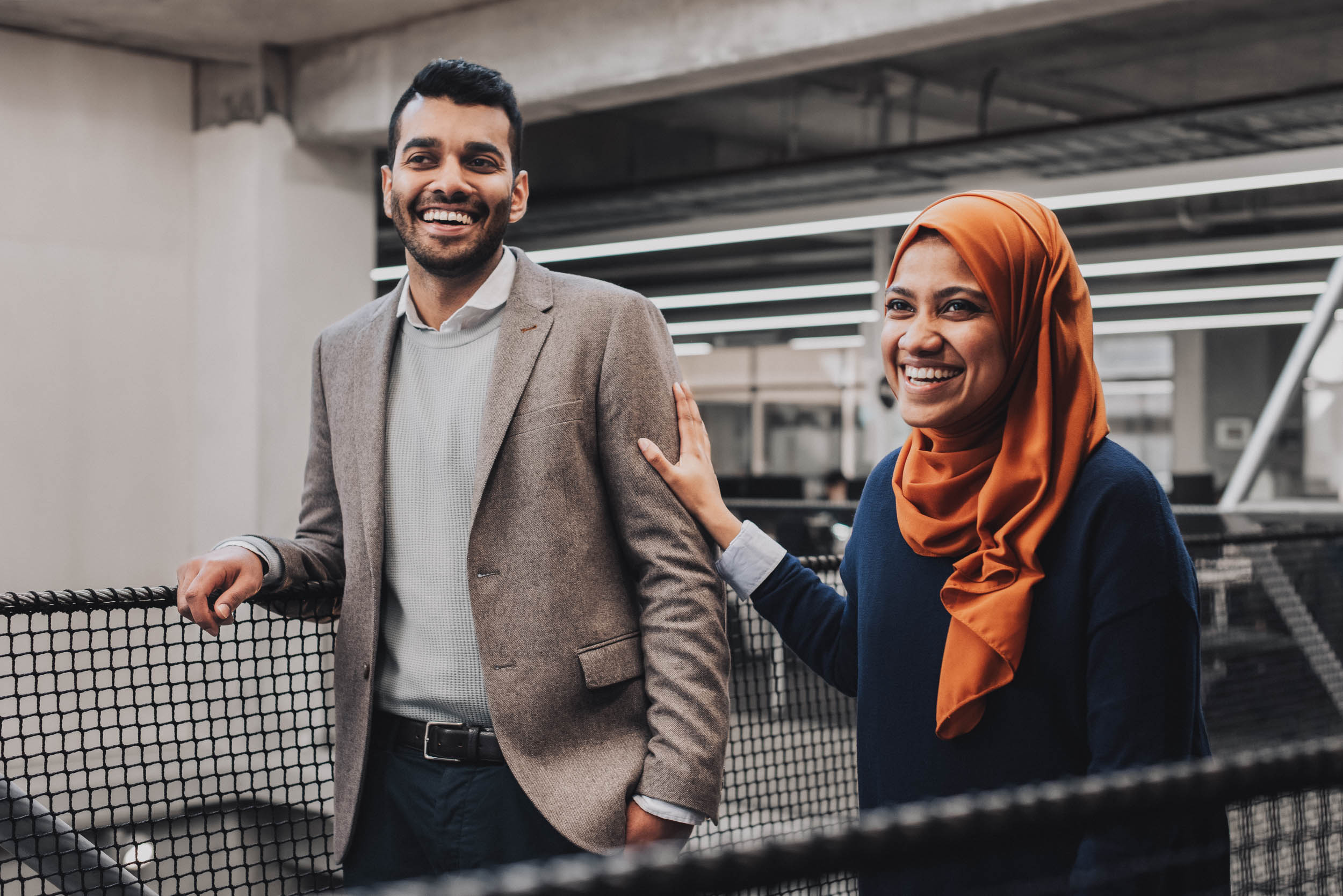Portrait of a smiling man in a blazer and a woman in a hijab, standing together indoors with natural lighting and neutral background.