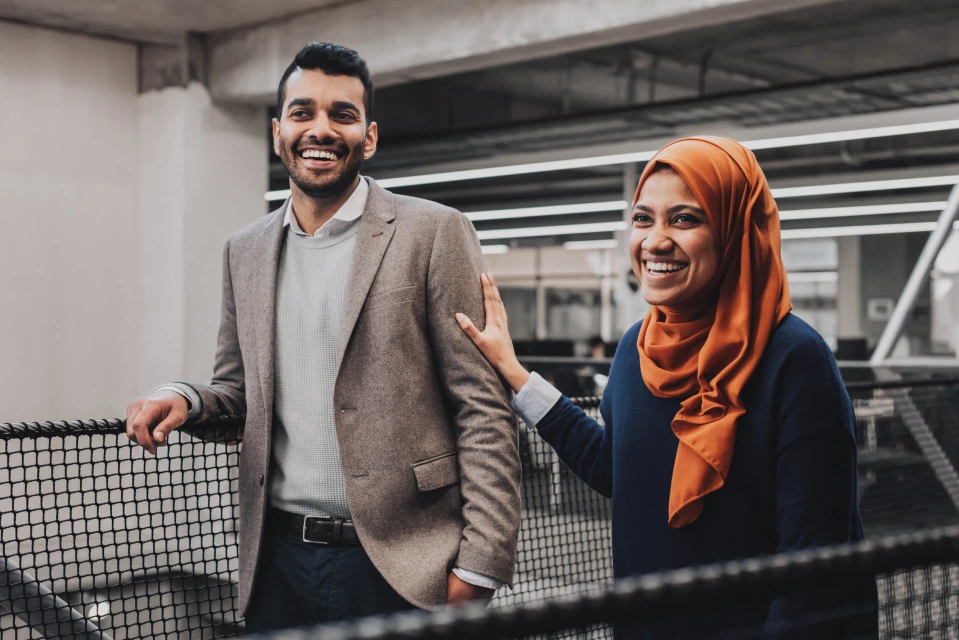 Portrait of a smiling man in a blazer and a woman in a hijab, standing together indoors with natural lighting and neutral background.