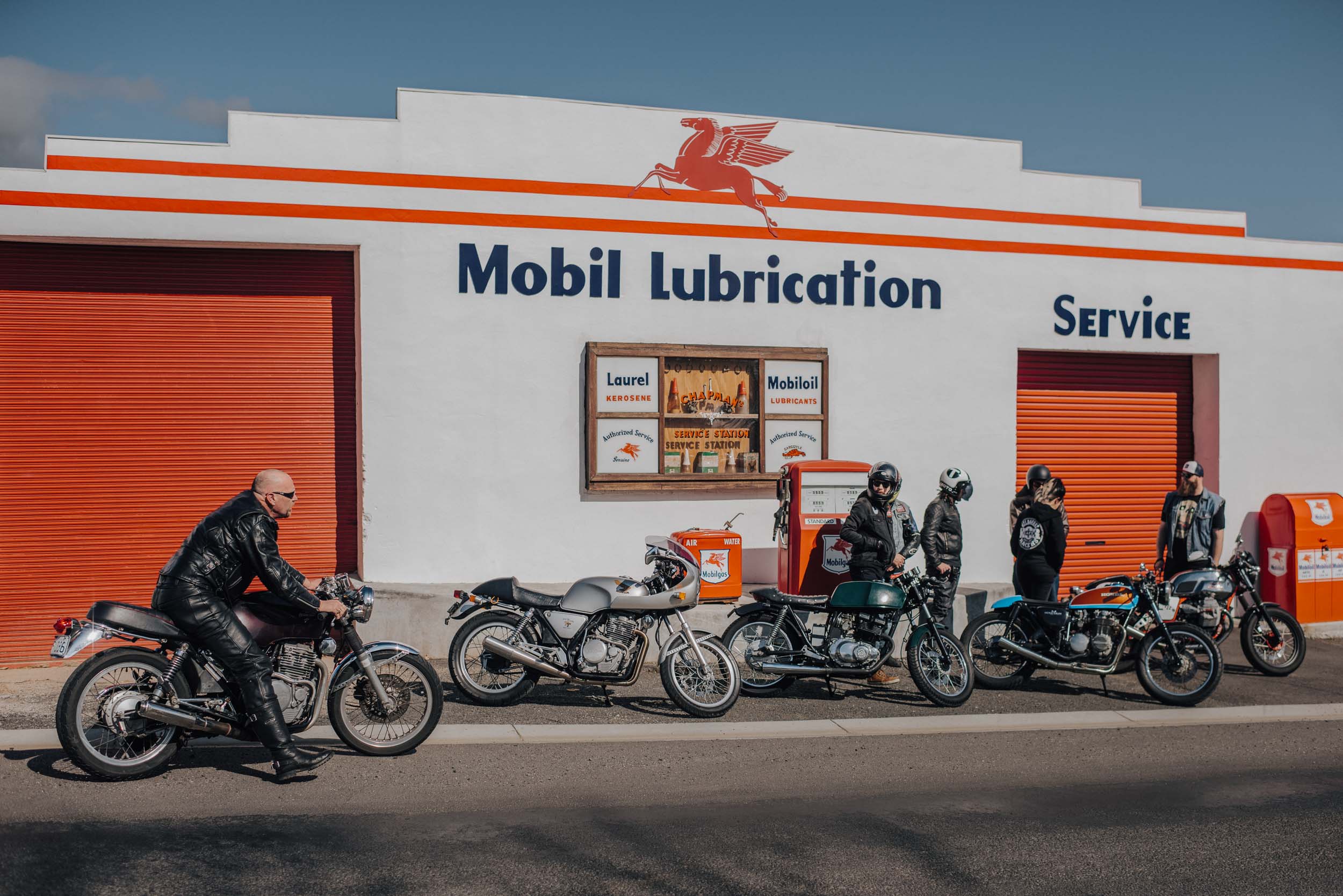 A vintage gas station scene with five motorcycles parked in front, men in riding gear, and a white building with orange accents.